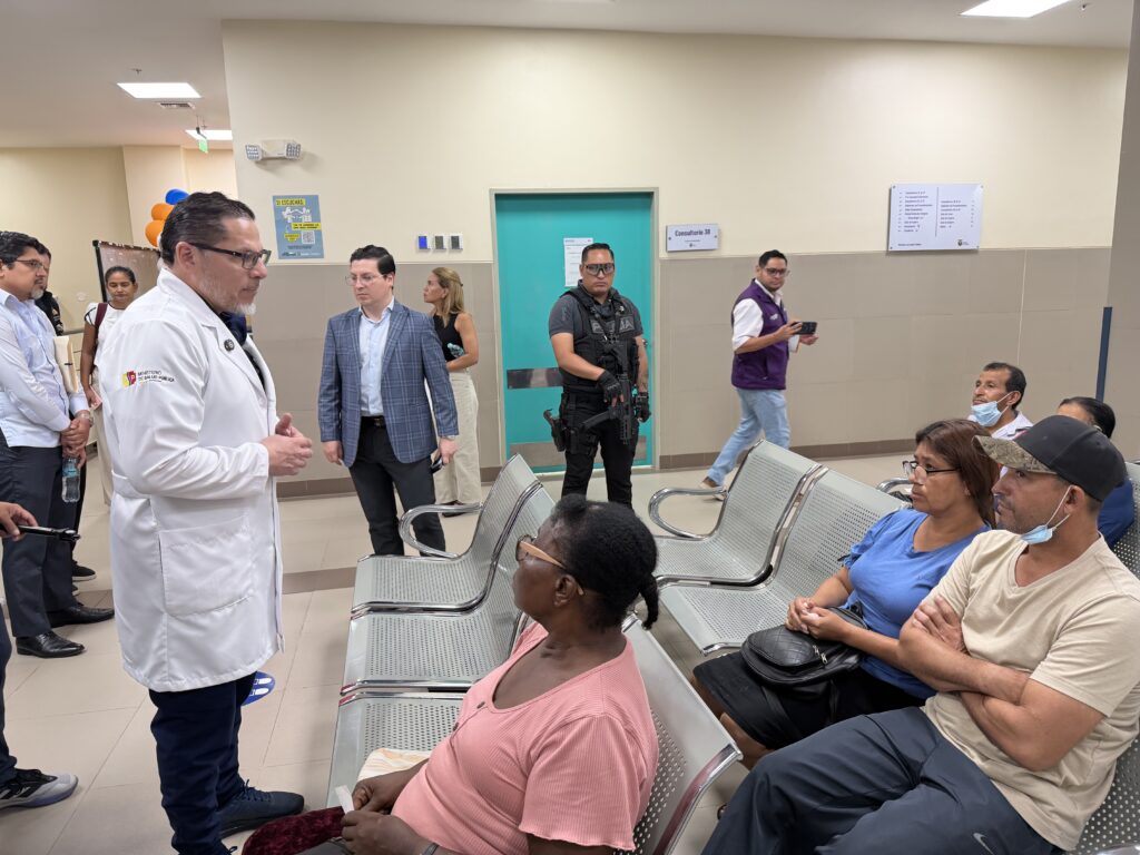 Representante del Ministerio de Salud conversa con usuarios en una sala de espera hospitalaria, junto a un afiche con código QR para denuncias.