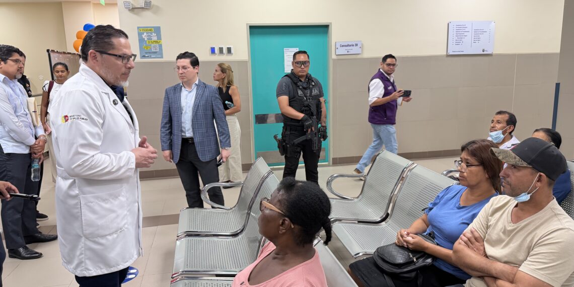 Representante del Ministerio de Salud conversa con usuarios en una sala de espera hospitalaria, junto a un afiche con código QR para denuncias.