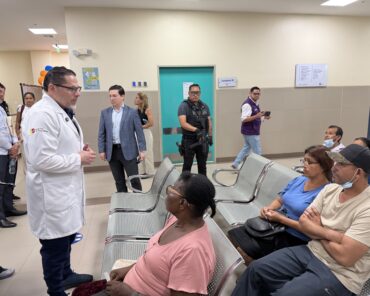 Representante del Ministerio de Salud conversa con usuarios en una sala de espera hospitalaria, junto a un afiche con código QR para denuncias.