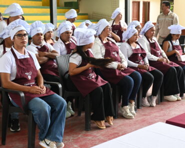 Participantes del curso de cocina en Santa Cecilia, sentados con gorros blancos y delantales granates durante la clausura.